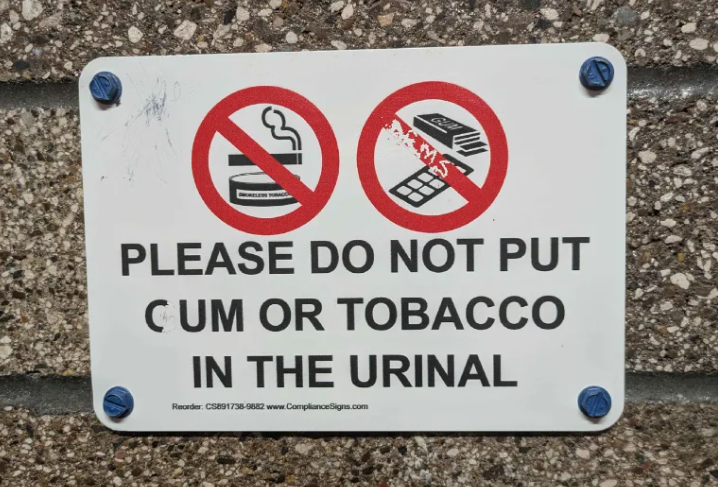 Pictured is a sign bolted to a brick wall that reads as follows: "Please do not put cum or tobacco in the urinal." The word "cum" on the sign shows signs of having read as "gum" before someone scrapped the "G" enough to turn it into a "C"